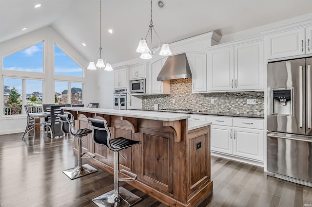 kitchen with white cabinetry, hanging light fixtures, wall chimney range hood, a kitchen island with sink, and appliances with stainless steel finishes