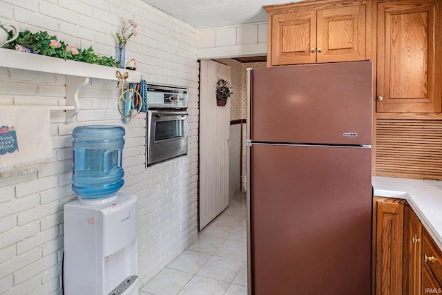 kitchen with wall oven, brick wall, light tile patterned floors, and stainless steel refrigerator