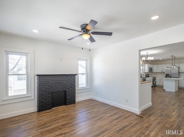 unfurnished living room with ceiling fan with notable chandelier, a brick fireplace, a wealth of natural light, and dark wood-type flooring