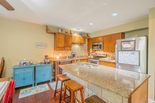 kitchen with appliances with stainless steel finishes, an island with sink, light stone countertops, dark hardwood / wood-style flooring, and sink