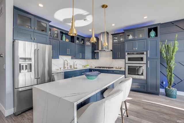 kitchen featuring stainless steel appliances, blue cabinets, wall chimney range hood, and pendant lighting