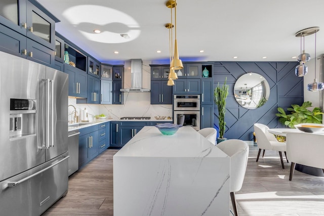 kitchen with stainless steel appliances, pendant lighting, light stone counters, wall chimney range hood, and blue cabinets