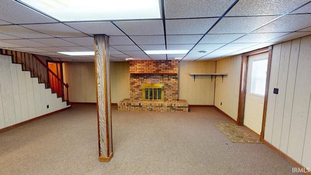 basement featuring light carpet, a brick fireplace, wood walls, and a drop ceiling