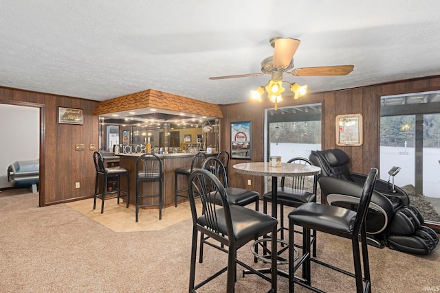 dining room with ceiling fan, wood walls, a textured ceiling, light carpet, and bar area