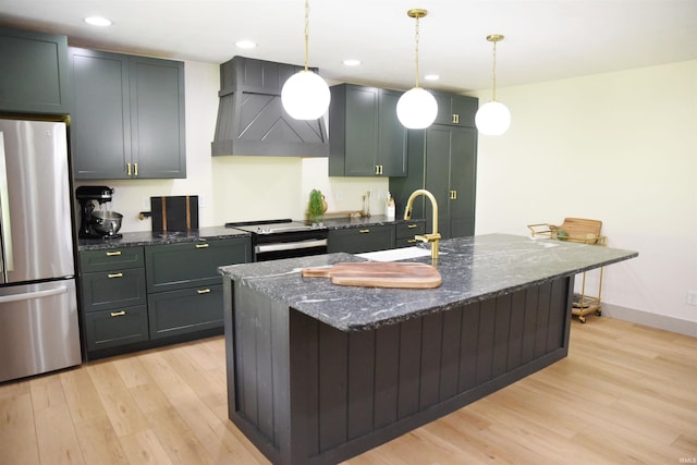 kitchen with pendant lighting, wall chimney exhaust hood, dark stone counters, an island with sink, and stainless steel fridge