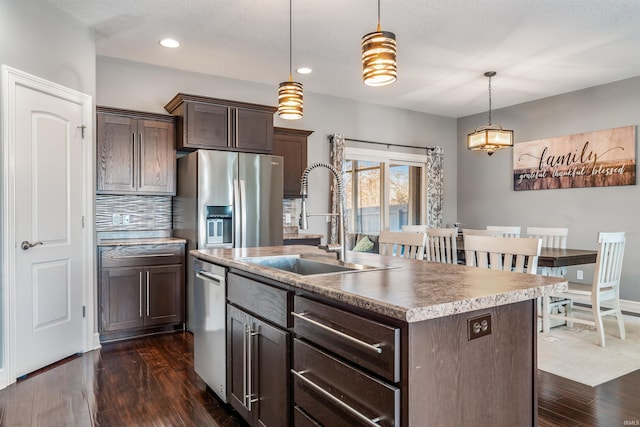 kitchen with an island with sink, stainless steel appliances, dark brown cabinets, hanging light fixtures, and sink