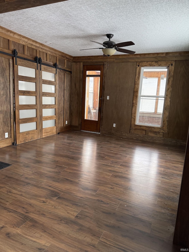 empty room with a barn door, wood walls, ceiling fan, dark hardwood / wood-style flooring, and a textured ceiling