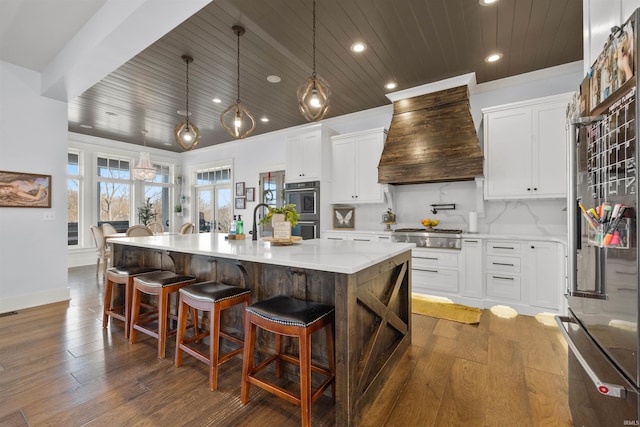 kitchen with a large island with sink, custom exhaust hood, white cabinetry, hanging light fixtures, and appliances with stainless steel finishes