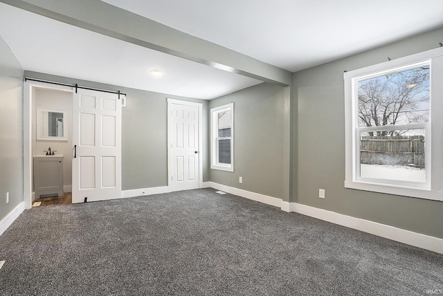 unfurnished bedroom with multiple windows, connected bathroom, dark carpet, and a barn door