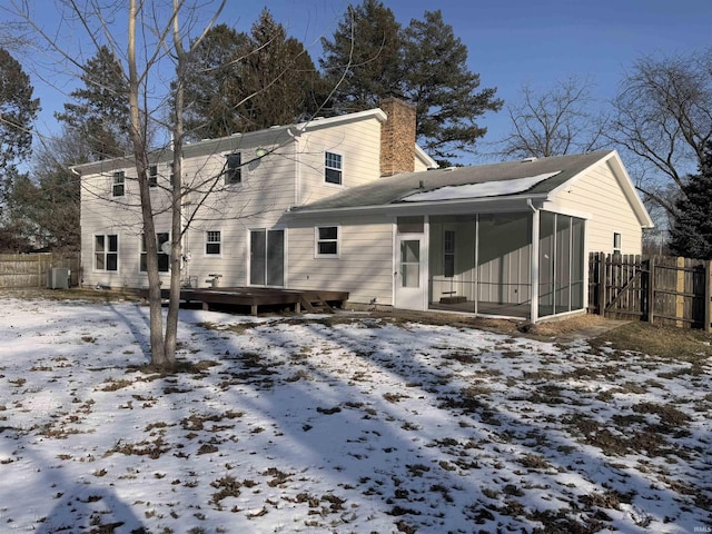 snow covered house featuring a sunroom, a chimney, fence, a deck, and central air condition unit