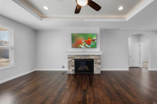 unfurnished living room with ceiling fan, a tray ceiling, a textured ceiling, dark hardwood / wood-style flooring, and a stone fireplace