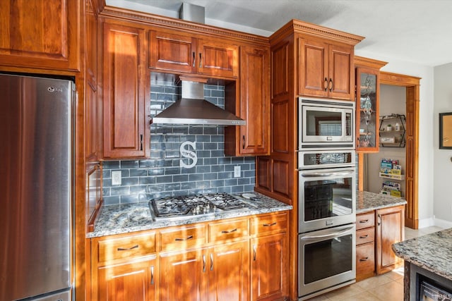 kitchen with backsplash, stainless steel appliances, light stone countertops, light tile patterned flooring, and wall chimney exhaust hood