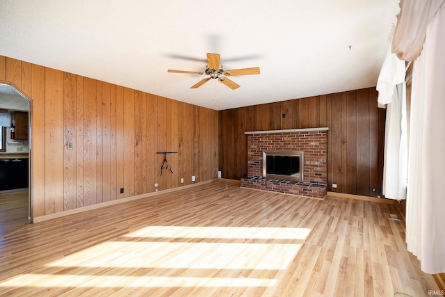 unfurnished living room featuring ceiling fan, wooden walls, a brick fireplace, and light wood-type flooring