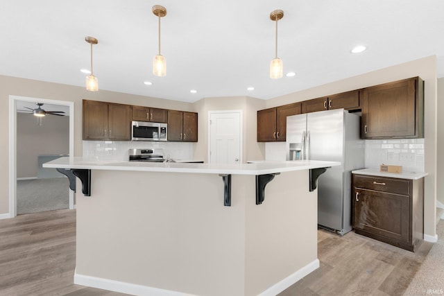 kitchen featuring appliances with stainless steel finishes, tasteful backsplash, an island with sink, hanging light fixtures, and dark brown cabinets