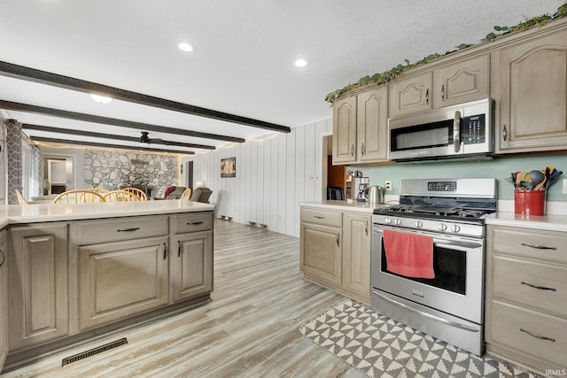 kitchen featuring stainless steel appliances, light brown cabinetry, a stone fireplace, beamed ceiling, and light wood-type flooring