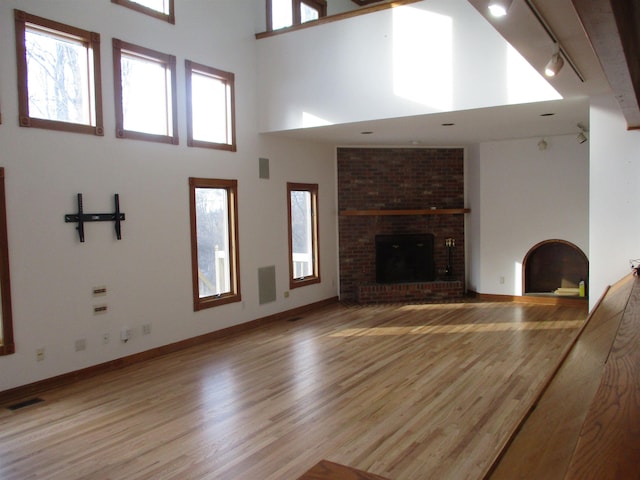 unfurnished living room featuring a brick fireplace, track lighting, a towering ceiling, a healthy amount of sunlight, and light hardwood / wood-style floors