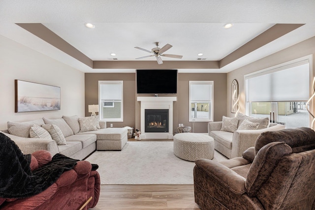 living room with ceiling fan, a fireplace, a raised ceiling, and light wood-type flooring