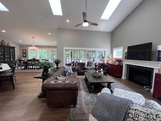 living room with ceiling fan with notable chandelier, high vaulted ceiling, a skylight, wood-type flooring, and a tiled fireplace