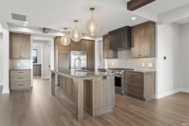 kitchen with pendant lighting, wall chimney range hood, a kitchen island with sink, beam ceiling, and premium appliances