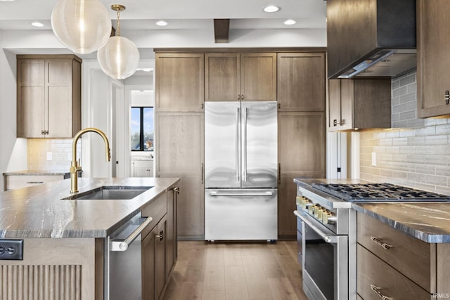 kitchen with sink, a center island with sink, premium appliances, dark stone counters, and wall chimney range hood