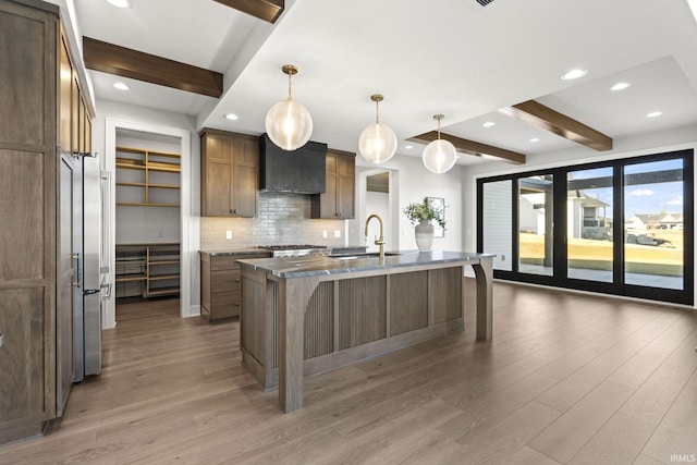 kitchen with light stone counters, tasteful backsplash, hanging light fixtures, an island with sink, and wall chimney range hood