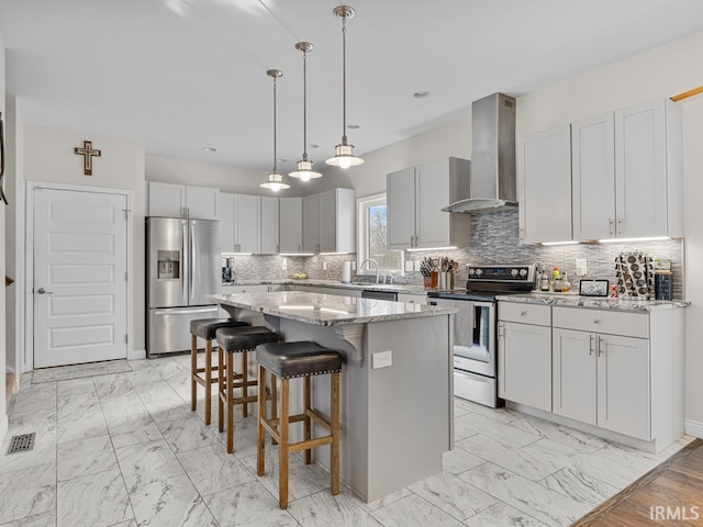 kitchen with wall chimney exhaust hood, light stone counters, a center island, hanging light fixtures, and appliances with stainless steel finishes