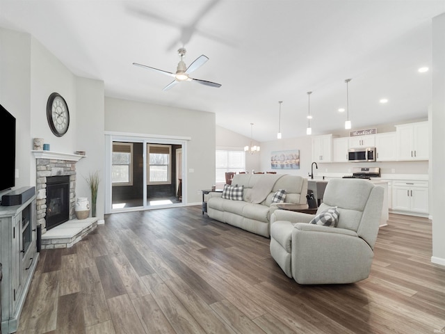 living room featuring lofted ceiling, sink, ceiling fan with notable chandelier, wood-type flooring, and a stone fireplace