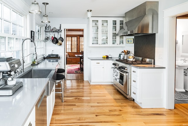 kitchen with wall chimney range hood, high end stainless steel range oven, white cabinetry, hanging light fixtures, and light wood-type flooring