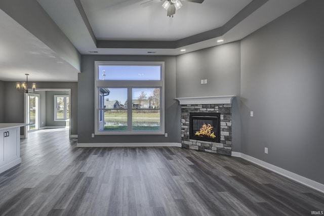 unfurnished living room with a stone fireplace, dark wood-type flooring, ceiling fan with notable chandelier, and a tray ceiling