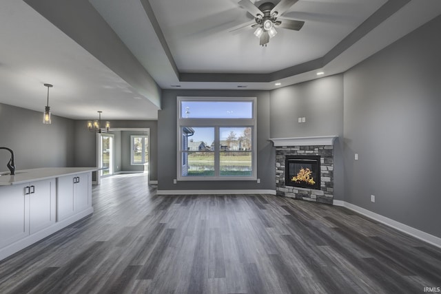 unfurnished living room featuring a stone fireplace, ceiling fan with notable chandelier, dark hardwood / wood-style flooring, and a tray ceiling