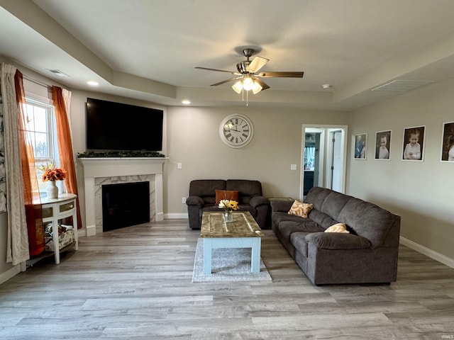 living room featuring a high end fireplace, light hardwood / wood-style floors, a raised ceiling, and ceiling fan