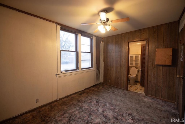 unfurnished bedroom with ceiling fan, carpet flooring, wooden walls, and ensuite bath