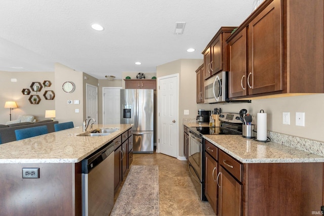 kitchen with sink, a textured ceiling, appliances with stainless steel finishes, light stone countertops, and a kitchen island with sink