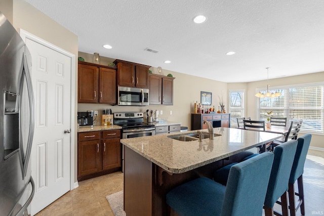 kitchen with sink, hanging light fixtures, stainless steel appliances, light stone countertops, and a kitchen island with sink