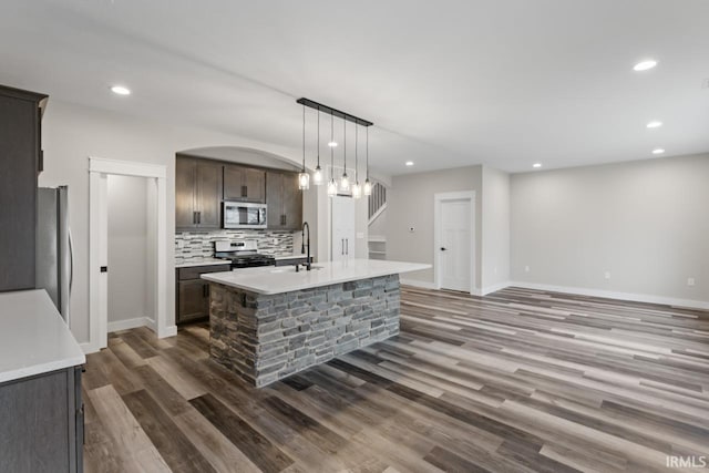 kitchen with appliances with stainless steel finishes, decorative backsplash, a kitchen island with sink, dark brown cabinetry, and dark wood-type flooring