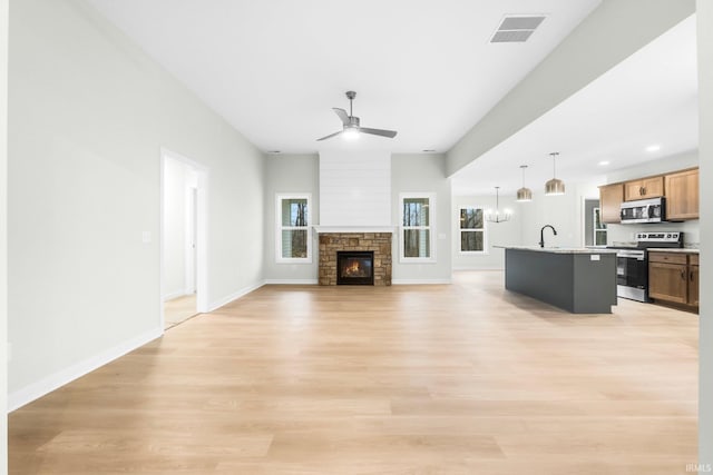 unfurnished living room featuring a stone fireplace, sink, ceiling fan with notable chandelier, and light hardwood / wood-style flooring