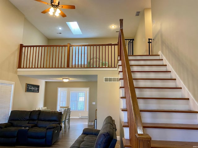 staircase with a towering ceiling, a skylight, a baseboard radiator, hardwood / wood-style flooring, and ceiling fan