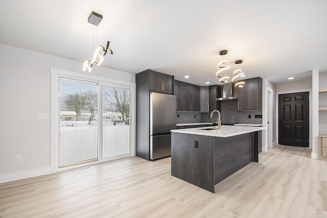 kitchen with dark brown cabinets, hanging light fixtures, wall chimney exhaust hood, stainless steel refrigerator, and a kitchen island with sink