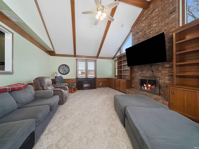 living area featuring ceiling fan, light colored carpet, a wainscoted wall, a brick fireplace, and beamed ceiling