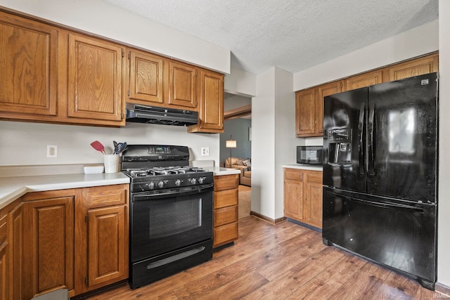 kitchen with light countertops, black appliances, brown cabinetry, light wood-type flooring, and under cabinet range hood