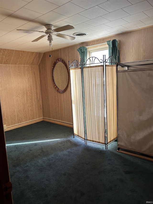 unfurnished room with wooden walls, visible vents, dark carpet, and a ceiling fan