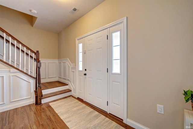 foyer entrance with a decorative wall, a wainscoted wall, wood finished floors, visible vents, and stairway