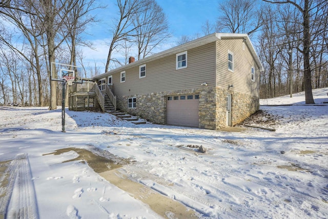 snow covered property featuring a garage, stone siding, a chimney, stairway, and a deck