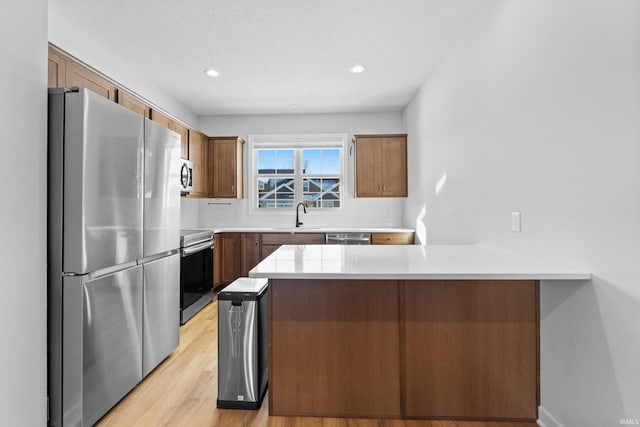 kitchen with brown cabinetry, appliances with stainless steel finishes, a peninsula, light countertops, and a sink