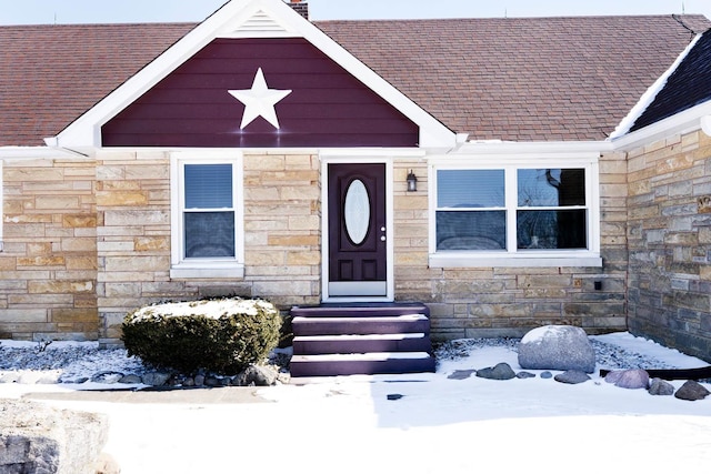 snow covered property entrance featuring a shingled roof and stone siding