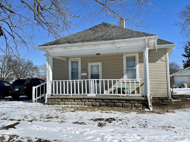 snow covered rear of property with covered porch, a shingled roof, and a chimney