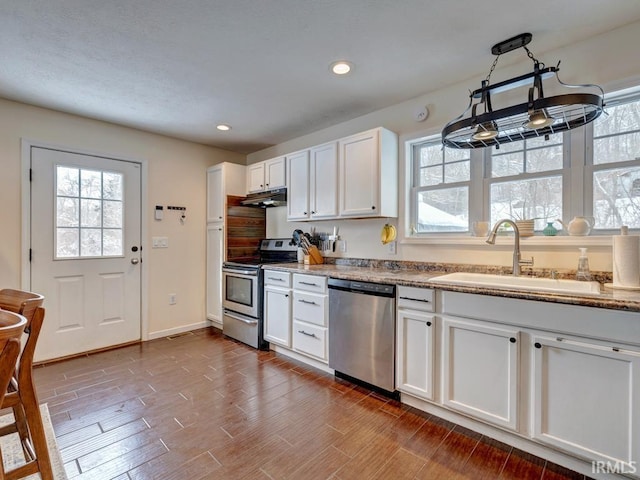 kitchen with under cabinet range hood, stainless steel appliances, a sink, white cabinets, and hanging light fixtures