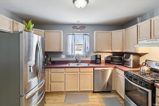 kitchen featuring dark countertops, light brown cabinetry, appliances with stainless steel finishes, a sink, and under cabinet range hood