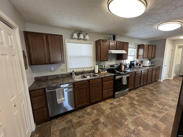 kitchen with dark brown cabinetry, under cabinet range hood, stainless steel appliances, a sink, and dark countertops