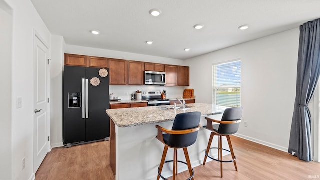 kitchen with light stone counters, stainless steel appliances, an island with sink, light wood-type flooring, and a kitchen breakfast bar